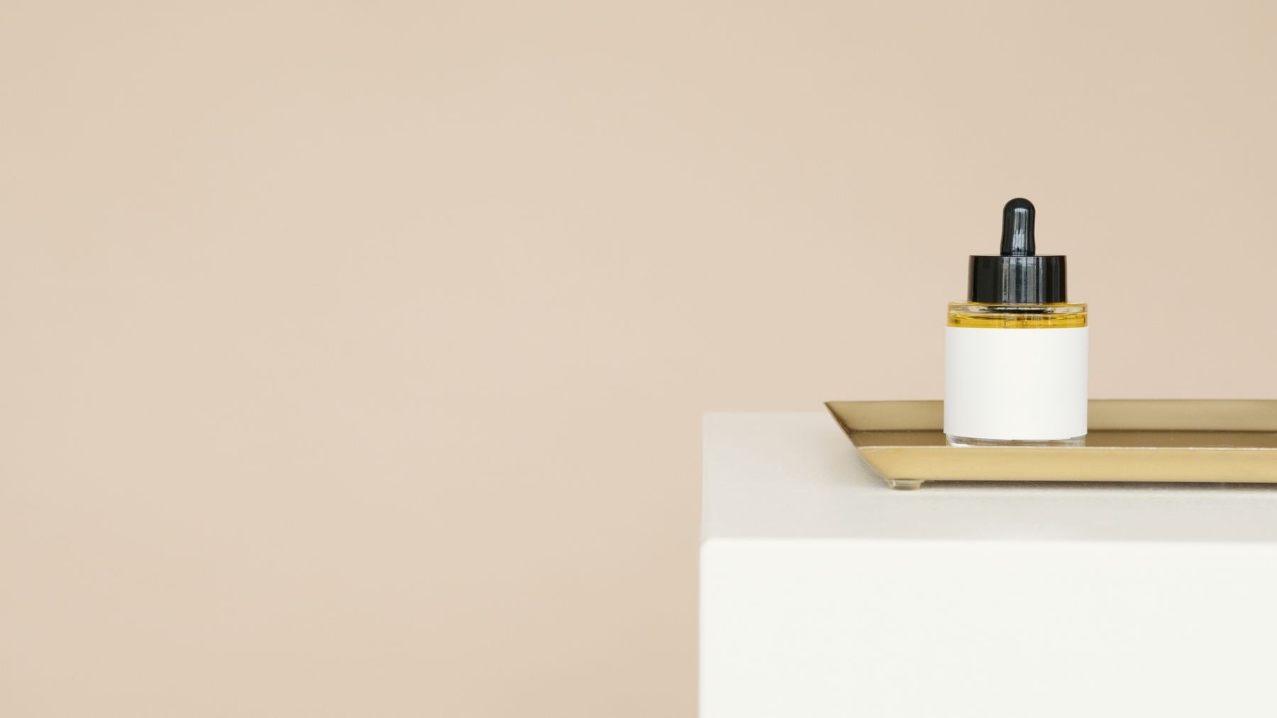 Glass dropper bottle on a brass tray at the edge of a white plinth against a warm neutral wall